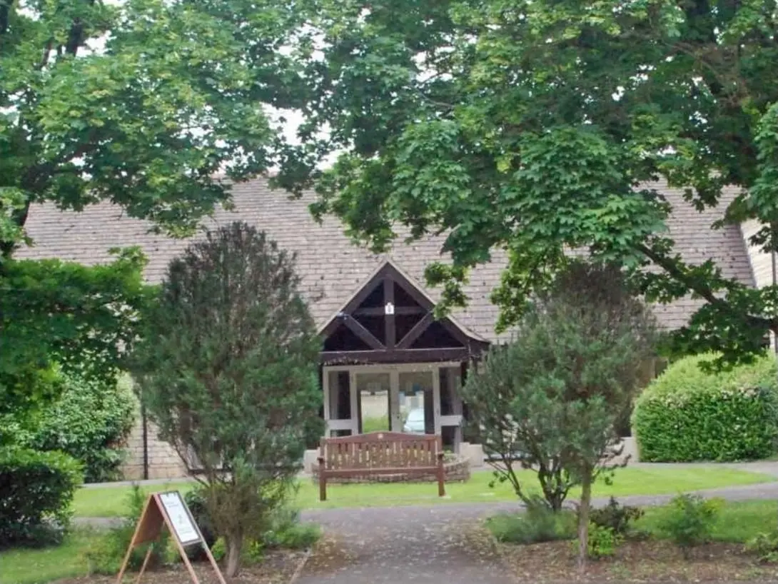 Bradwell Village Hall seen through tall trees, with a timber porch and a bench on the lawn in front