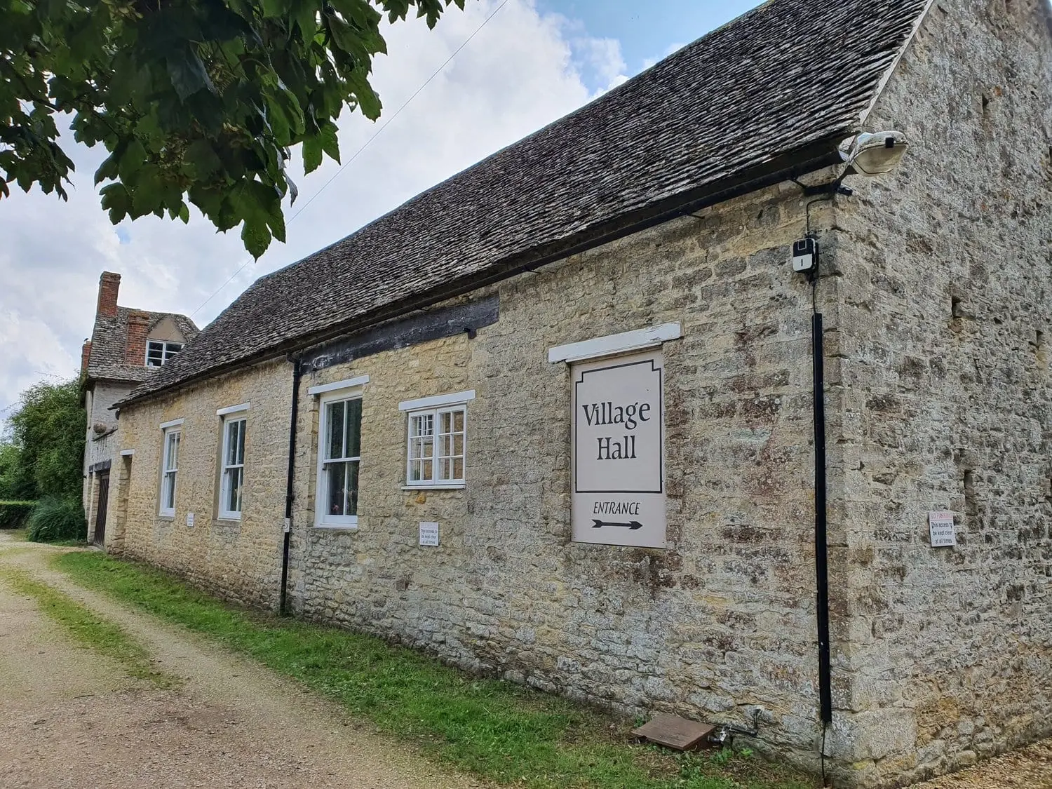 Ducklington Village Hall, a long Cotswold stone hall with a stone-tiled roof and a Village Hall entrance sign